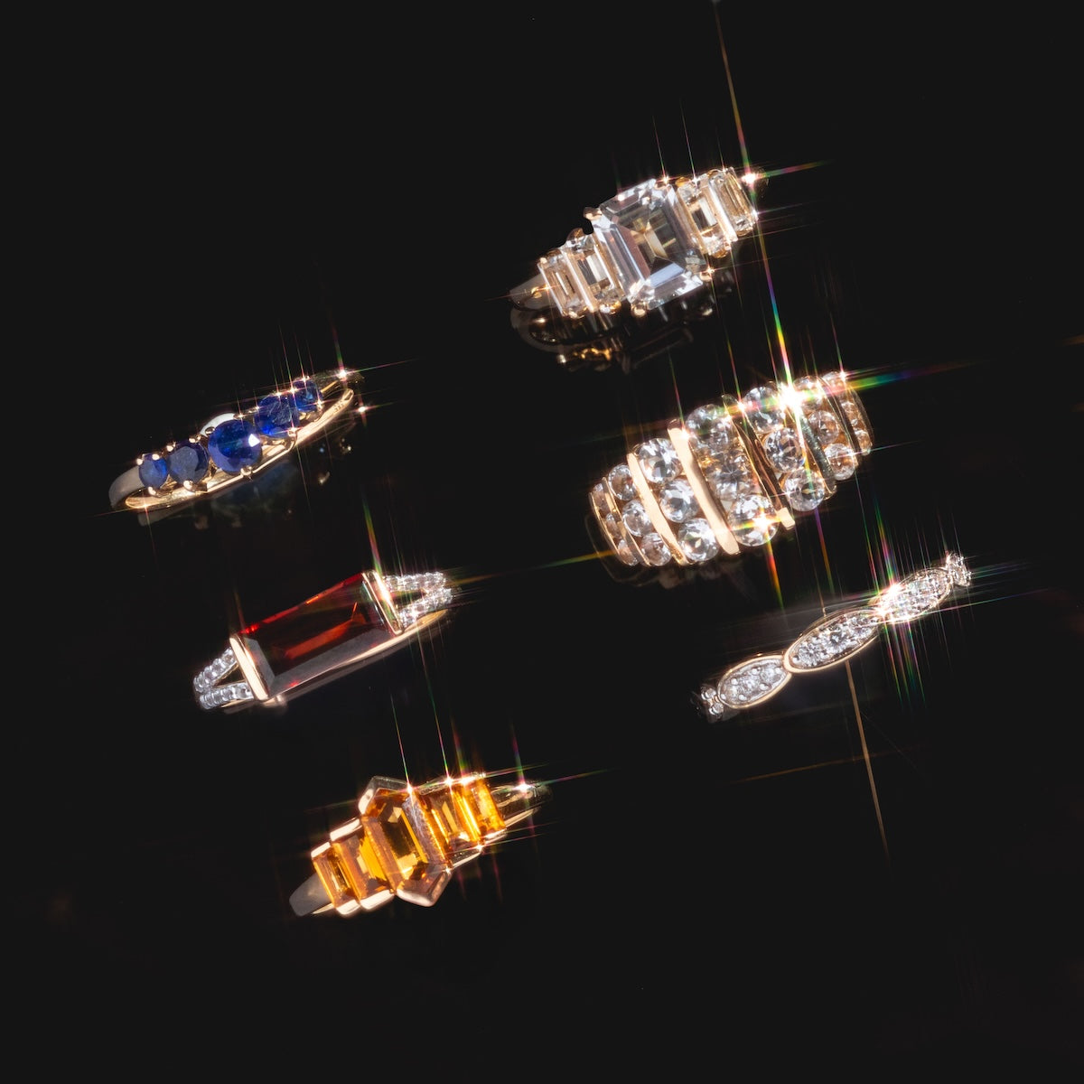 Six assorted gemstone and diamond rings, including the 14k Graduated Citrine Deco Ring, are displayed on a glossy black surface. The collection features blue, red, and yellow stones with diamonds in intricate gold settings.