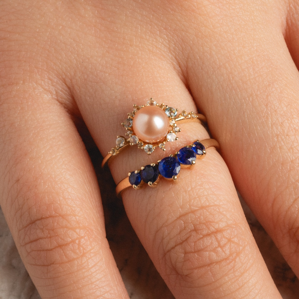 A close-up of a hand wearing two gold rings: a 14k yellow gold pearl and diamond ring, and the 14k Blue Sapphire Arc Nesting Ring featuring five sapphires set in a row.