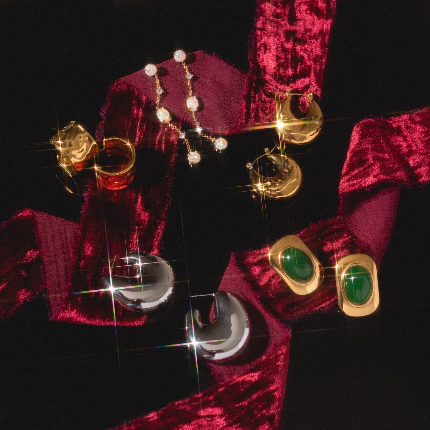 Ashley Earrings in gold, silver, and green gemstone styles displayed on a dark surface with a burgundy velvet ribbon, sparkling reflections highlighting the jewelry.
