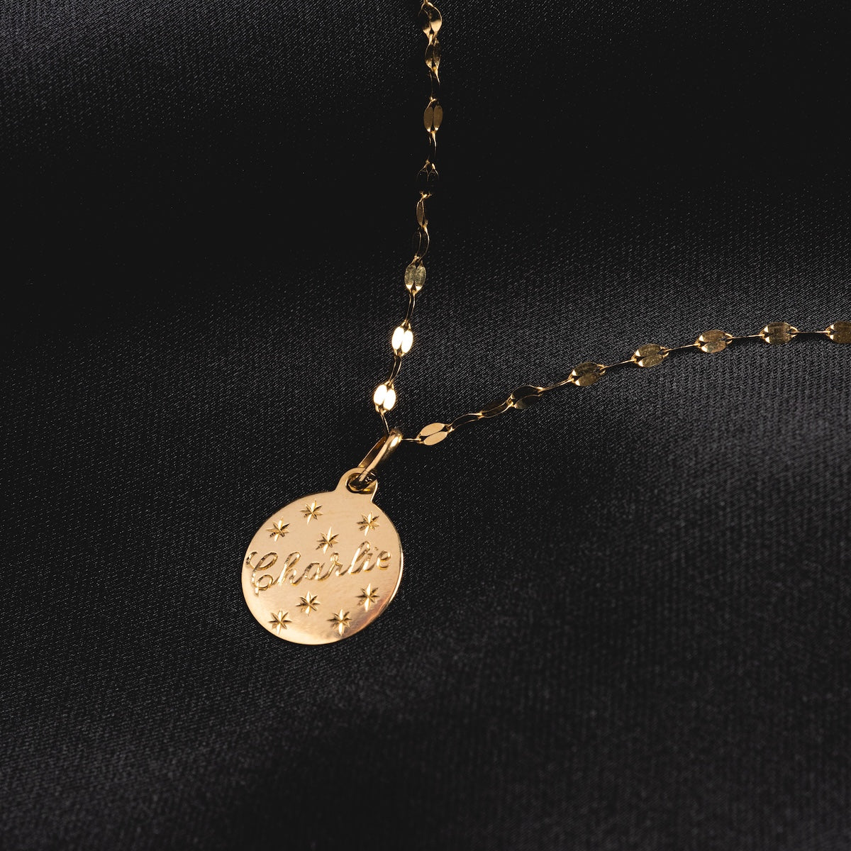 The 14k Written in the Stars Customizable Name Charm is a personalized gold necklace with a round pendant engraved with “Charlie” and star designs, beautifully presented on a black fabric background.