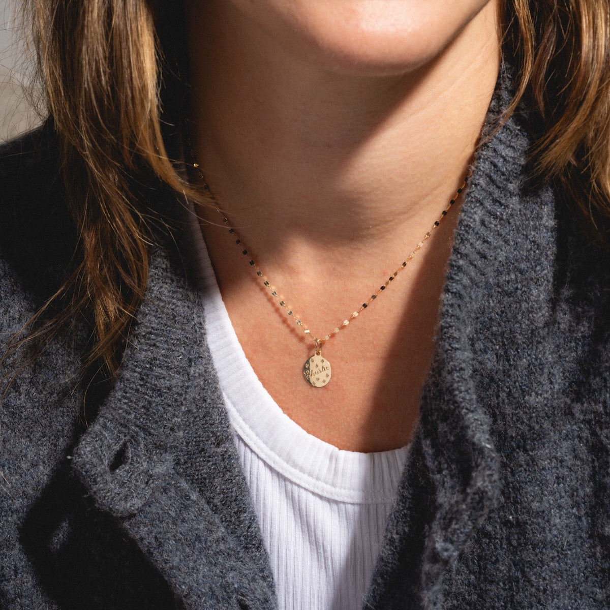 A person wears a dark gray textured cardigan over a white ribbed shirt, accessorized with the 14k Written in the Stars Customizable Name Charm—a delicate gold necklace engraved with dots.