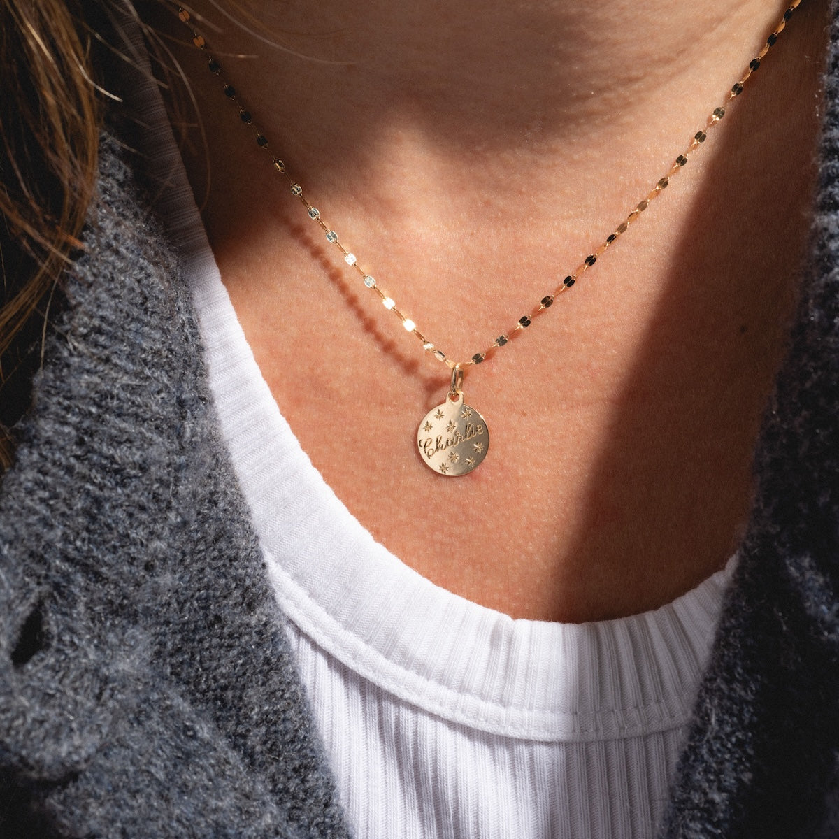 A person wears the 14k Written in the Stars Customizable Name Charm, a gold necklace with a round engraved pendant, over a white pleated shirt and dark textured sweater. The elegant jewelry features partially readable cursive text.