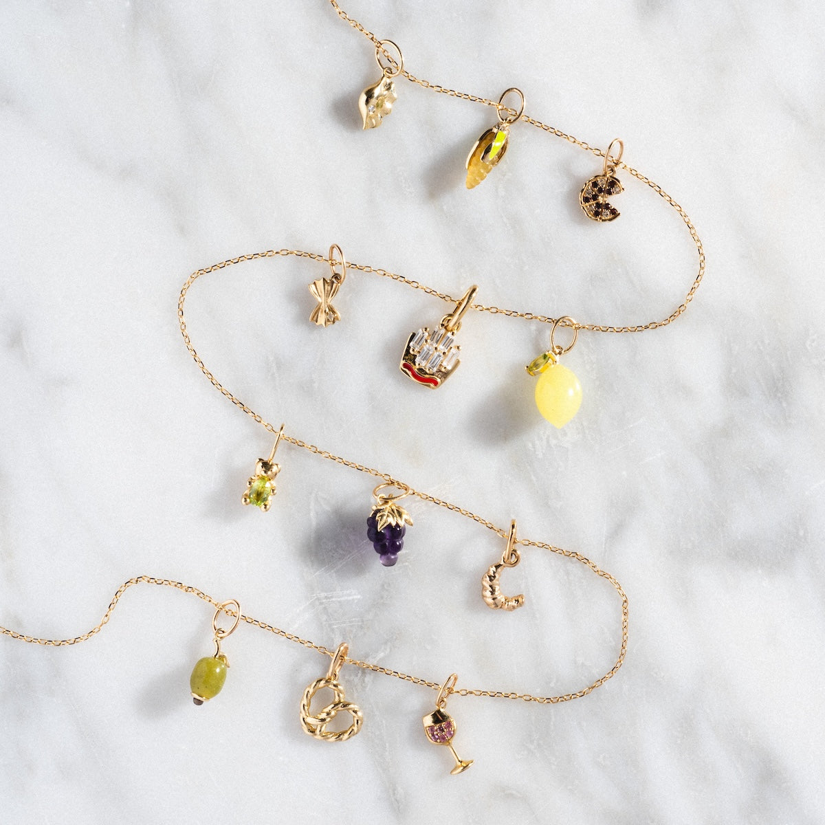 The Solid Gold Foodie Charm features three delicate 9k yellow gold chains with vibrant solid gold charms—pretzel, lemon, grapes, cocktail, cheese, and more—ideal for a playful necklace. Shown on a white marble surface.