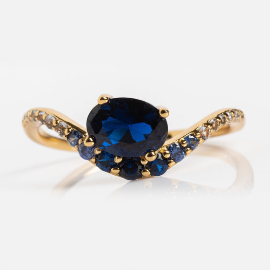 The Main Character Floating Ombre Birthstone Ring showcases an oval deep blue gemstone at the center, accented by smaller blue stones and diamonds on a delicate twisting band—a stunning piece of birthstone jewelry.