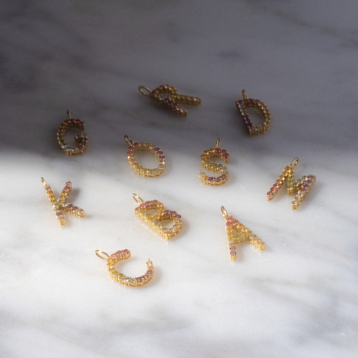 Nine Solid Gold Pastel Gemstone Letter Charms, featuring the letters C, O, S, M, R, D, K, B, and A with pastel gemstones, are arranged on a marble surface in soft natural light and delicate shadows.