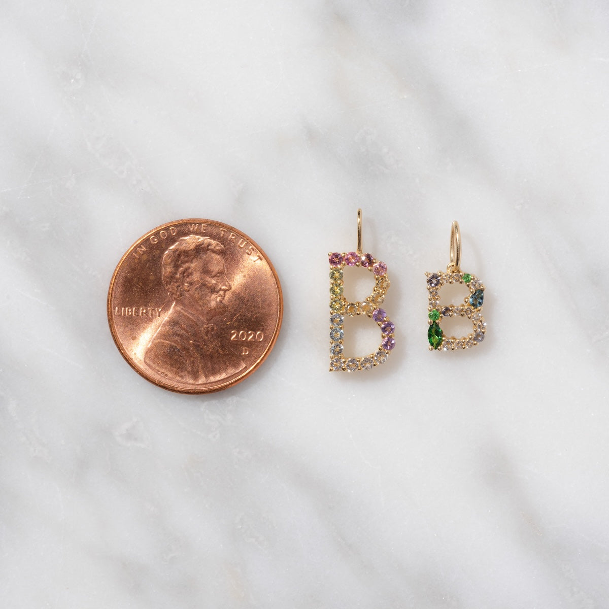 A 2020 U.S. penny beside two Solid Gold Pastel Gemstone Letter Charm hoop earrings, each shaped like the letter B and decorated with pastel gemstone charms, displayed on a white marble surface.