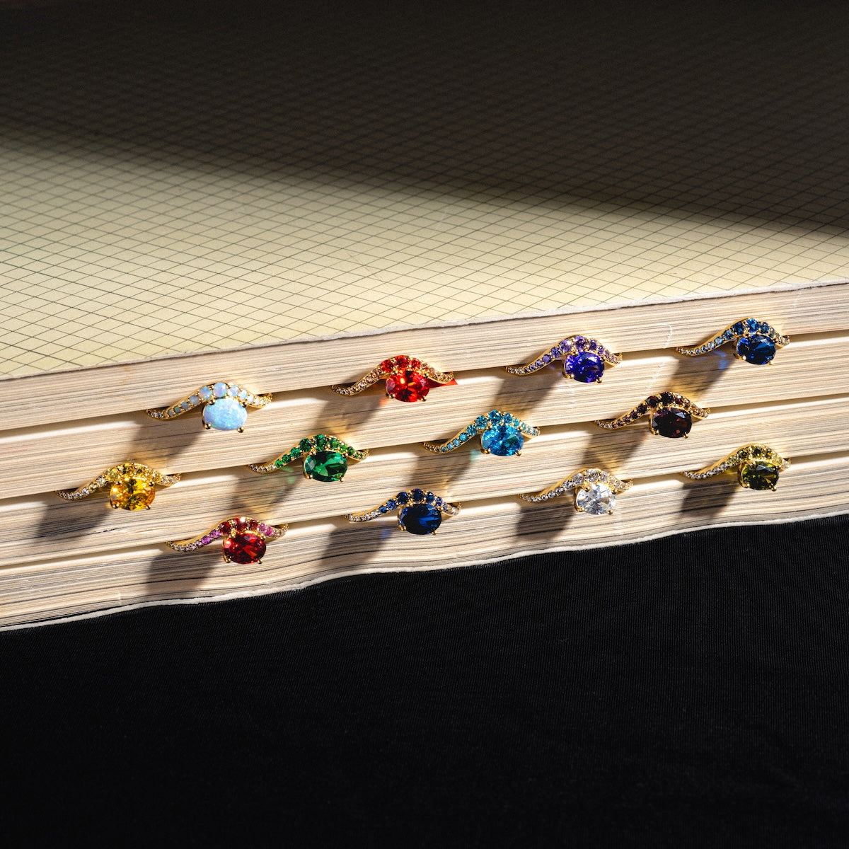 The Main Character Floating Ombre Birthstone Ring shines among fourteen gold rings with colorful gemstones, all neatly arranged on stacked grid-lined paper set against a black background.
