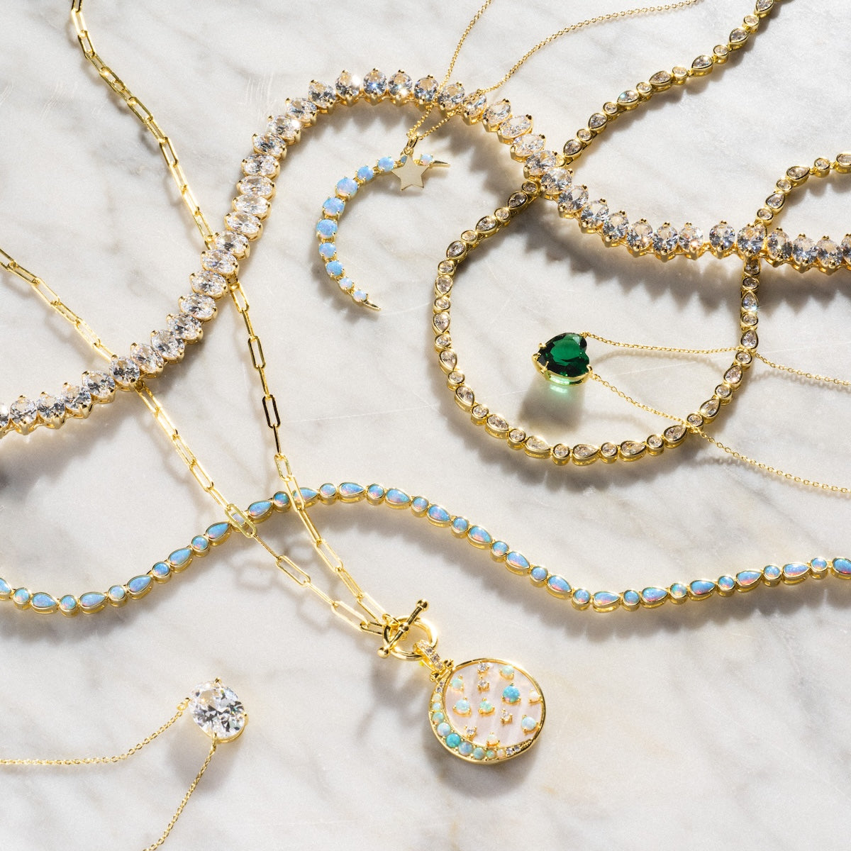 The Allison Emerald Heart Pendant Necklace, featuring a vibrant green gemstone, is displayed on light marble among gold and jeweled necklaces, bracelets, and an opal-and-diamond pendant, highlighting sparkling gems.