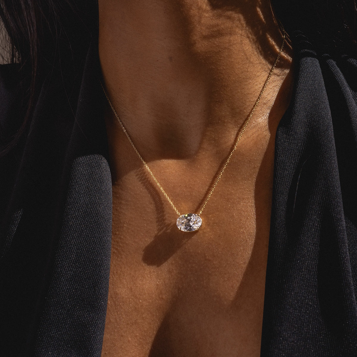 A close-up of a woman's neck wearing the Hello Gorgeous Necklace, a delicate 18k yellow gold chain with a large oval solitaire pendant, paired with a dark deep V-neck outfit.