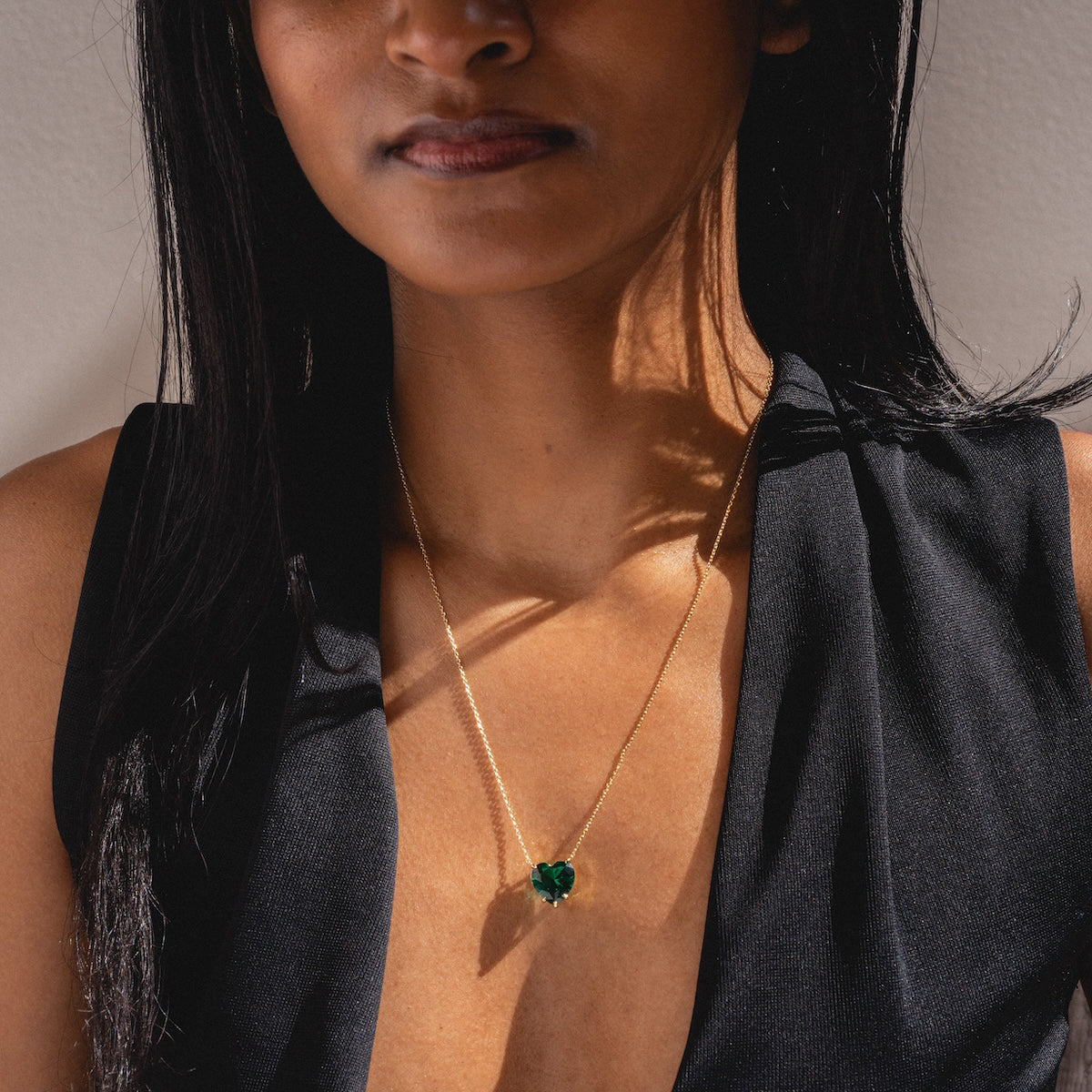 A woman wears a sleeveless black top and The Allison Emerald Heart Pendant Necklace, standing against a light background with sunlight creating shadows.