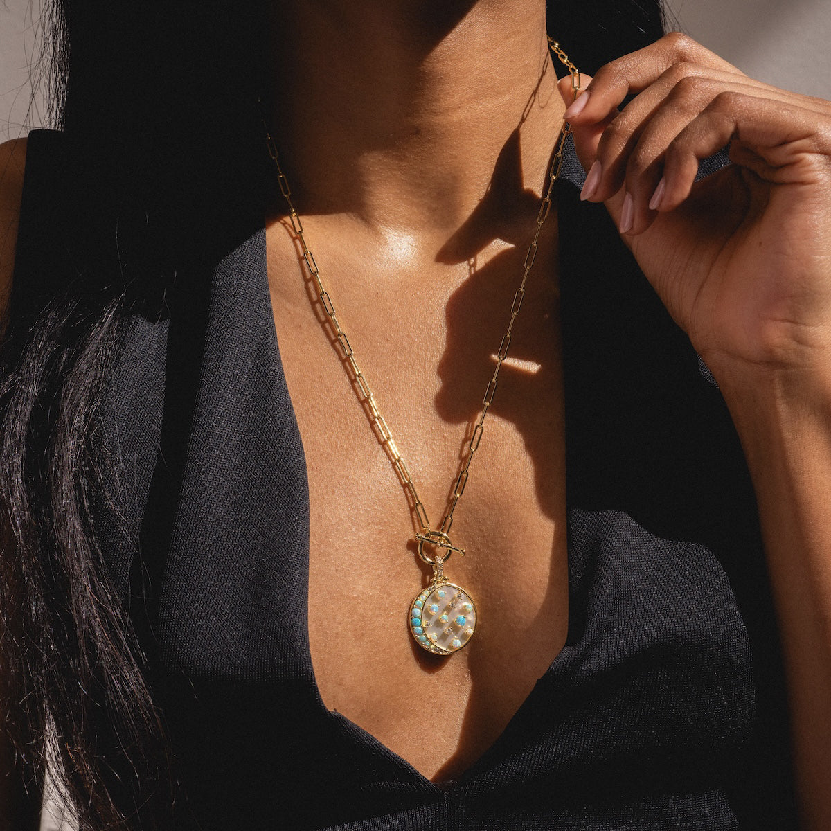 A woman in a deep v-neck black top models the Sweet Dreams Samantha Chain Necklace Sample, a chunky gold chain with an 18k yellow plated pendant featuring a round simulated opal centerpiece accented by small stones. Sunlight highlights the piece near her neck.