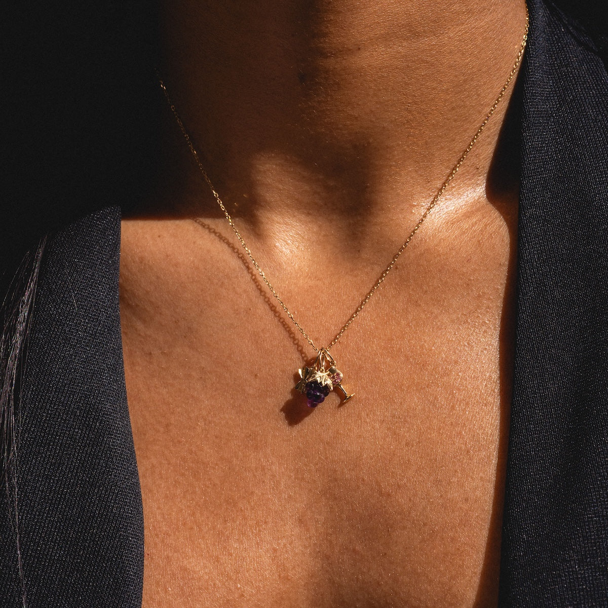 A close-up of a person wearing the Solid Gold Foodie Charm necklace in 9k yellow gold with a small, cluster-shaped pendant and purple gemstone, paired with a dark, open-collared top.