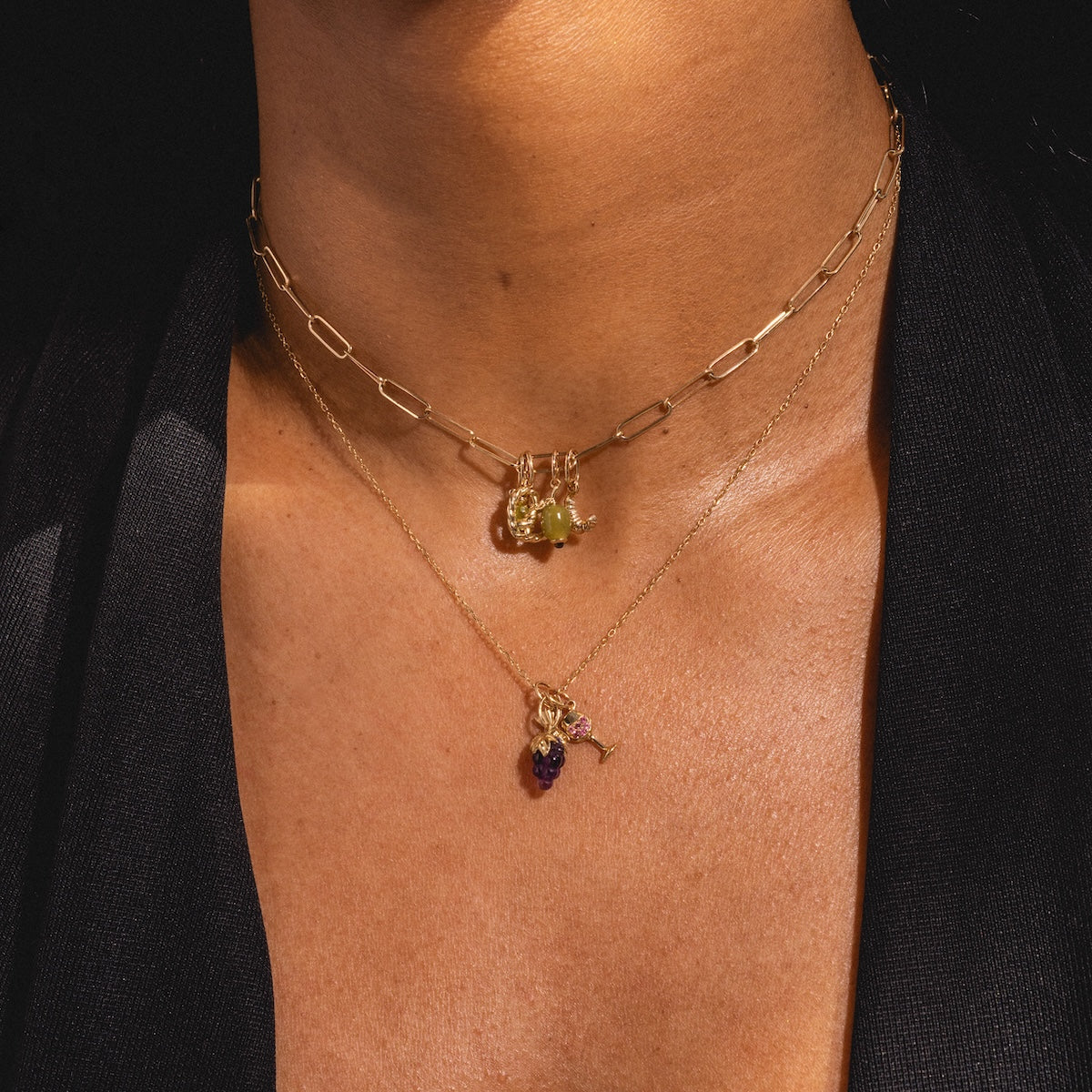 A close-up of a person layering two gold necklaces—a 9k yellow gold paperclip chain with green gemstone charm and a fine chain featuring the Solid Gold Foodie Charm in purple—over a dark top.
