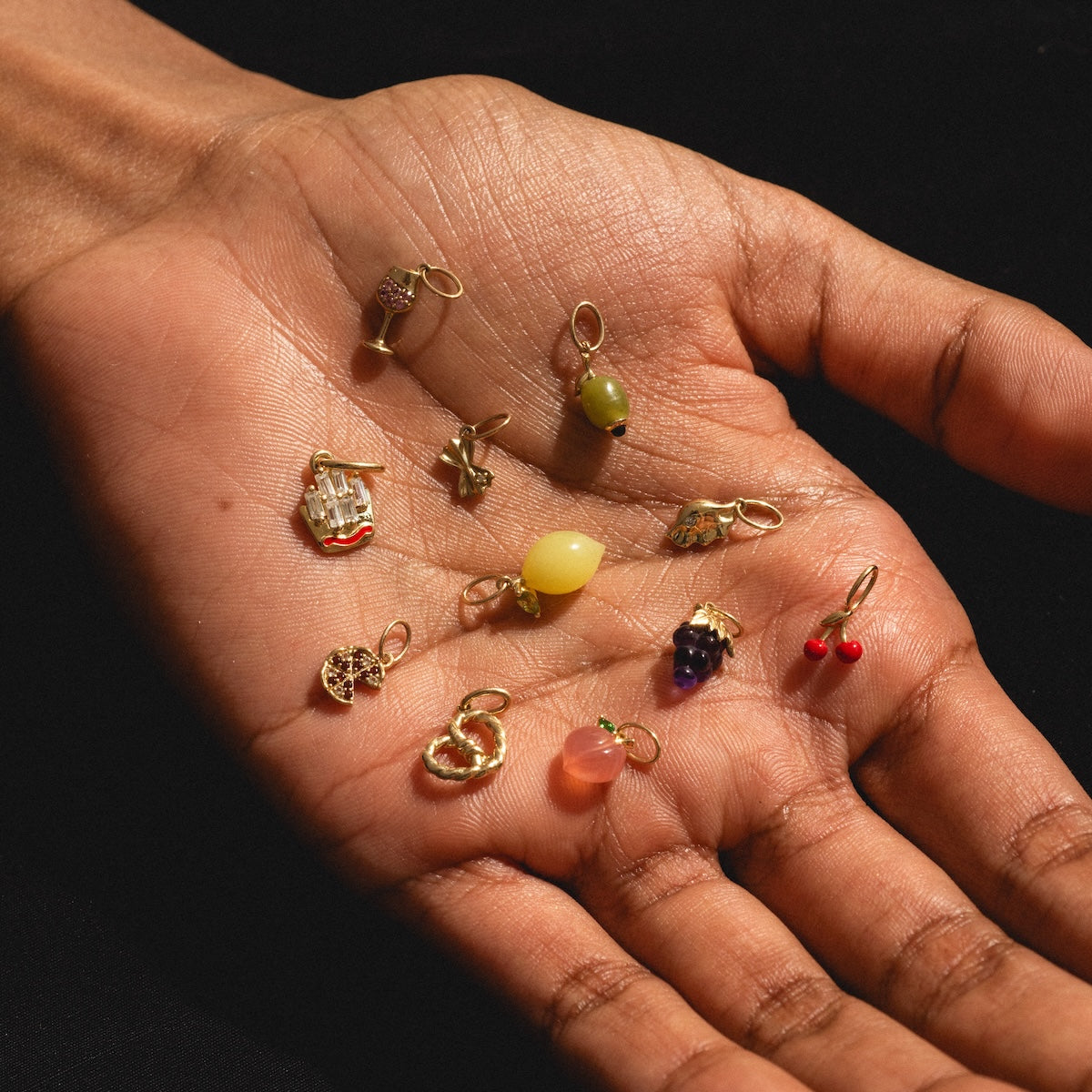 A hand displays nine Solid Gold Foodie Charms—each shaped like colorful fruits or foods with 9k yellow gold loops—against a dark background.