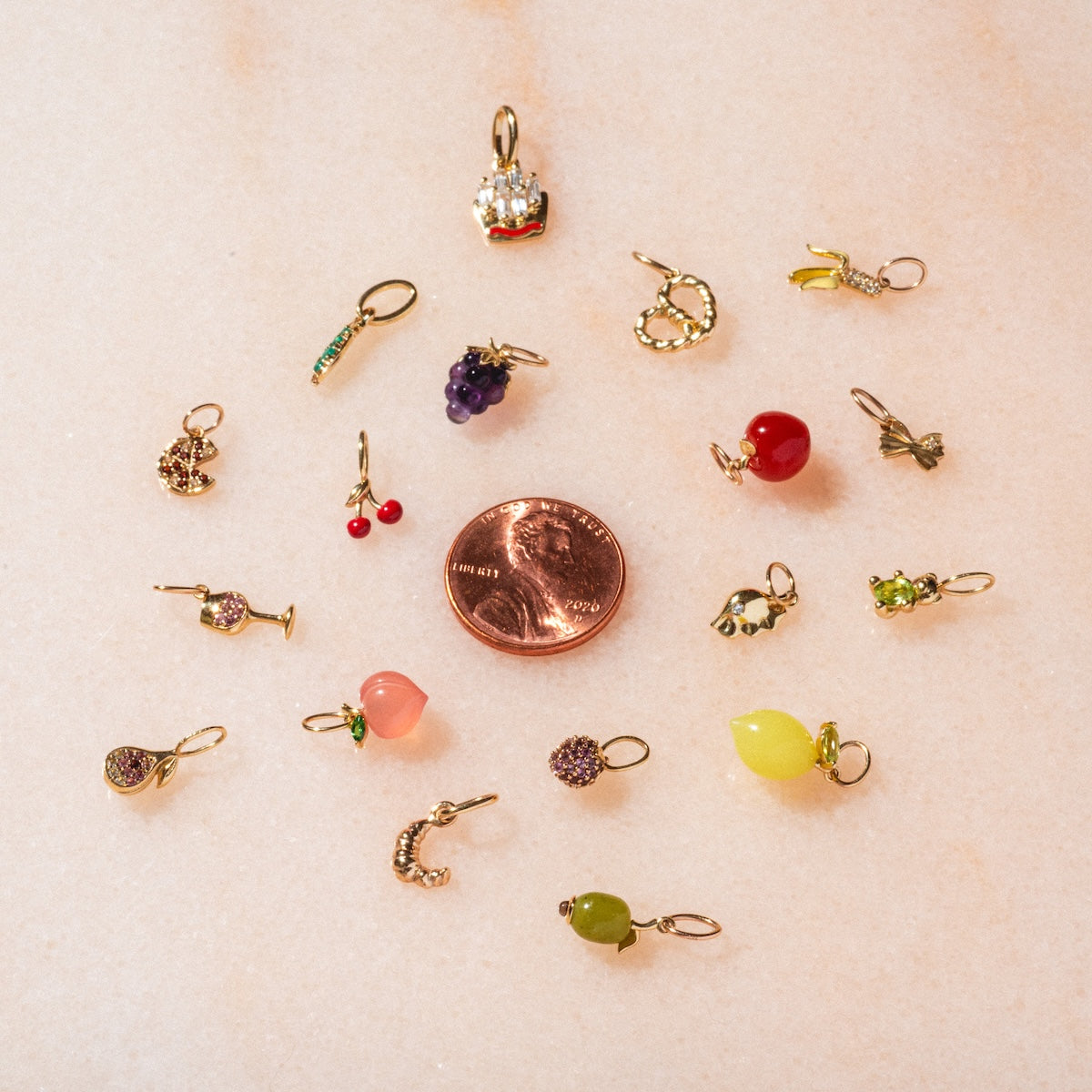 The Solid Gold Foodie Charm features small, colorful fruit and veggie shapes arranged around a penny on pale pink—ideal for playful necklaces or pairing with 9k yellow gold.