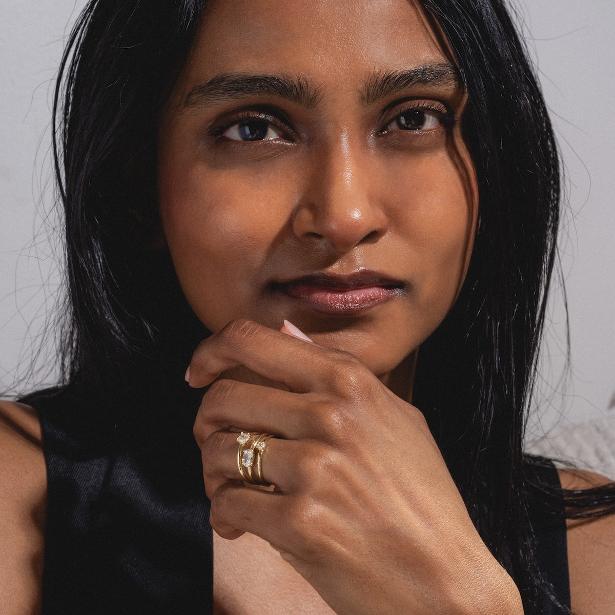 A woman with long dark hair gazes at the camera, resting her chin on her hand. She wears a sleeveless black top and the Nina Stacked Ring Sample Size 8, an 18k yellow gold plated band with simulated diamond accents. The background is softly lit.