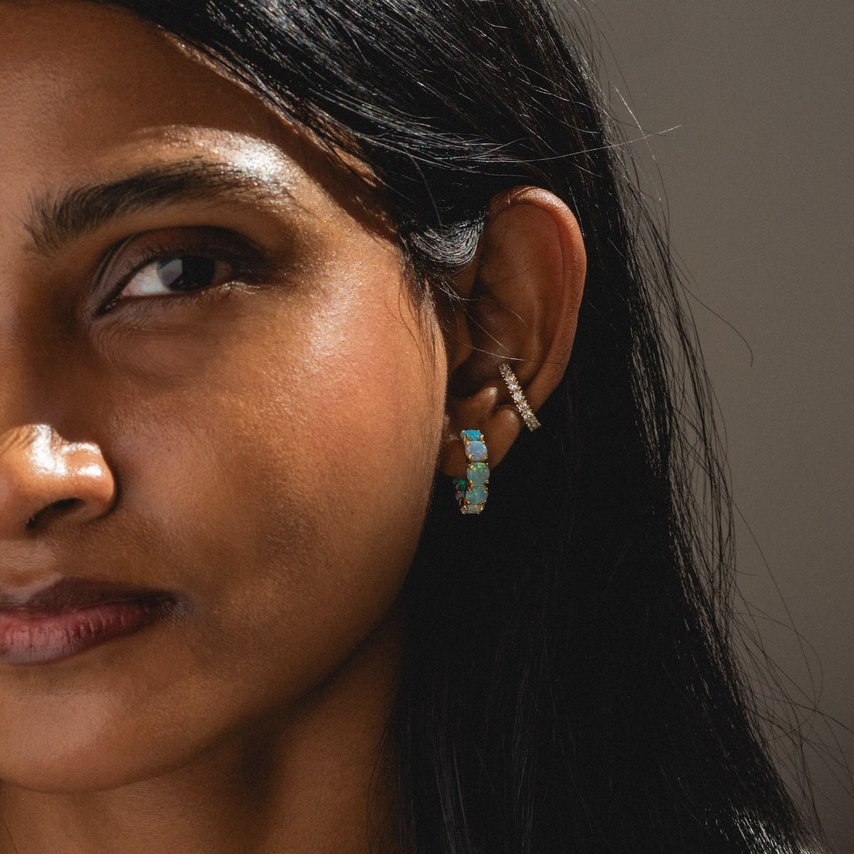 Close-up of a womans face, featuring her left ear with the Oh She Fancy Ombre Hoops—18k yellow gold plated turquoise and silver hoops—and a smaller silver textured cuff. She has long dark hair and brown skin.