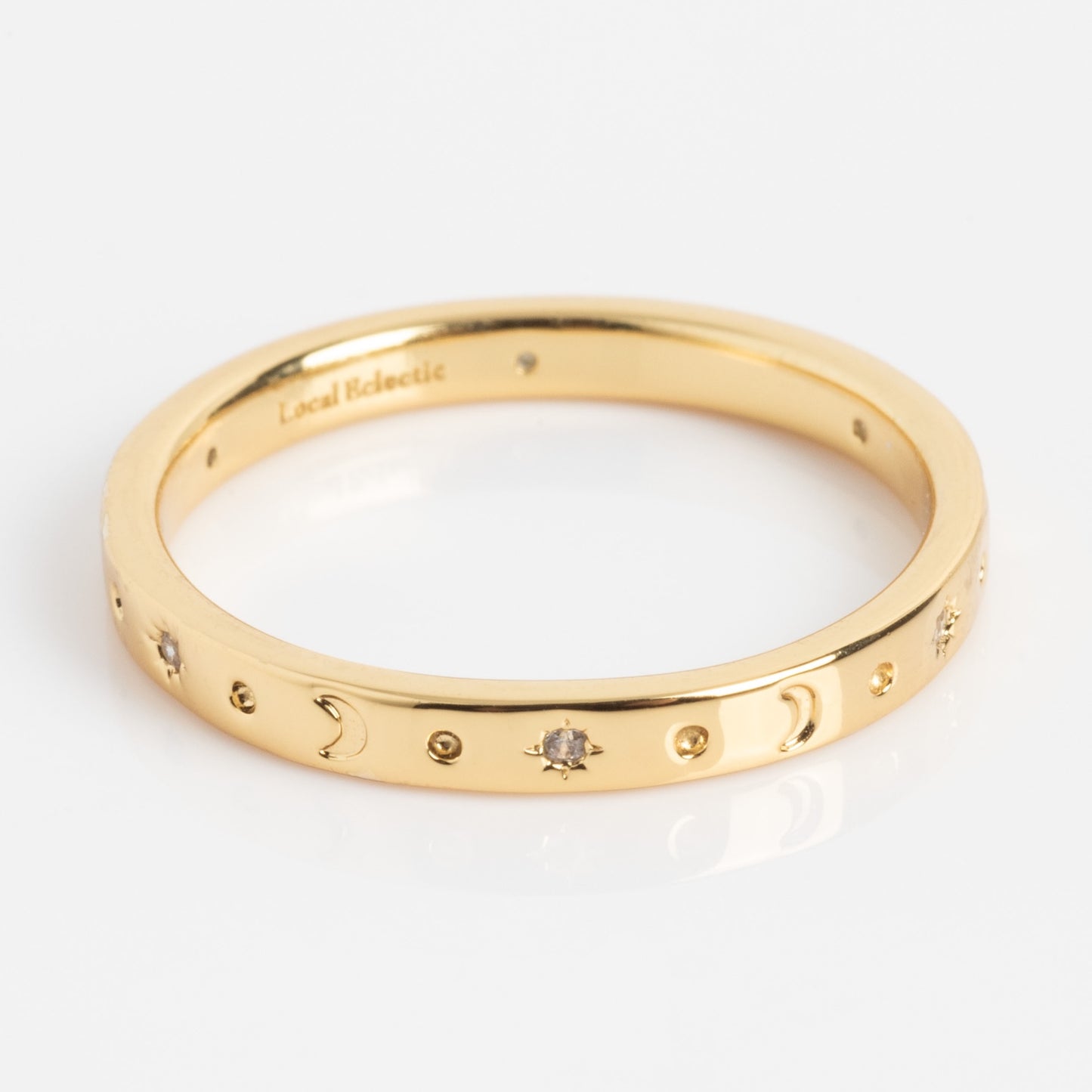 The Moon and Stars Stacking Ring is a thin gold band engraved with crescent moons, stars, and small gemstone accents spaced evenly around it, shown on a white background.