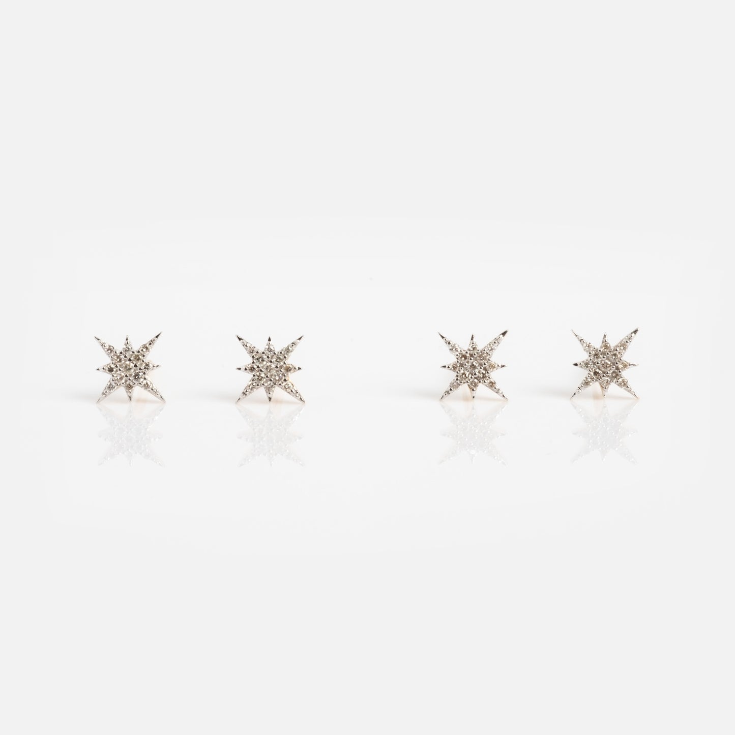 Four Solid Gold Diamond Starlight Studs, inspired by celestial designs and set with sparkling diamonds, are arranged in a row on a white background with a subtle reflection beneath them.