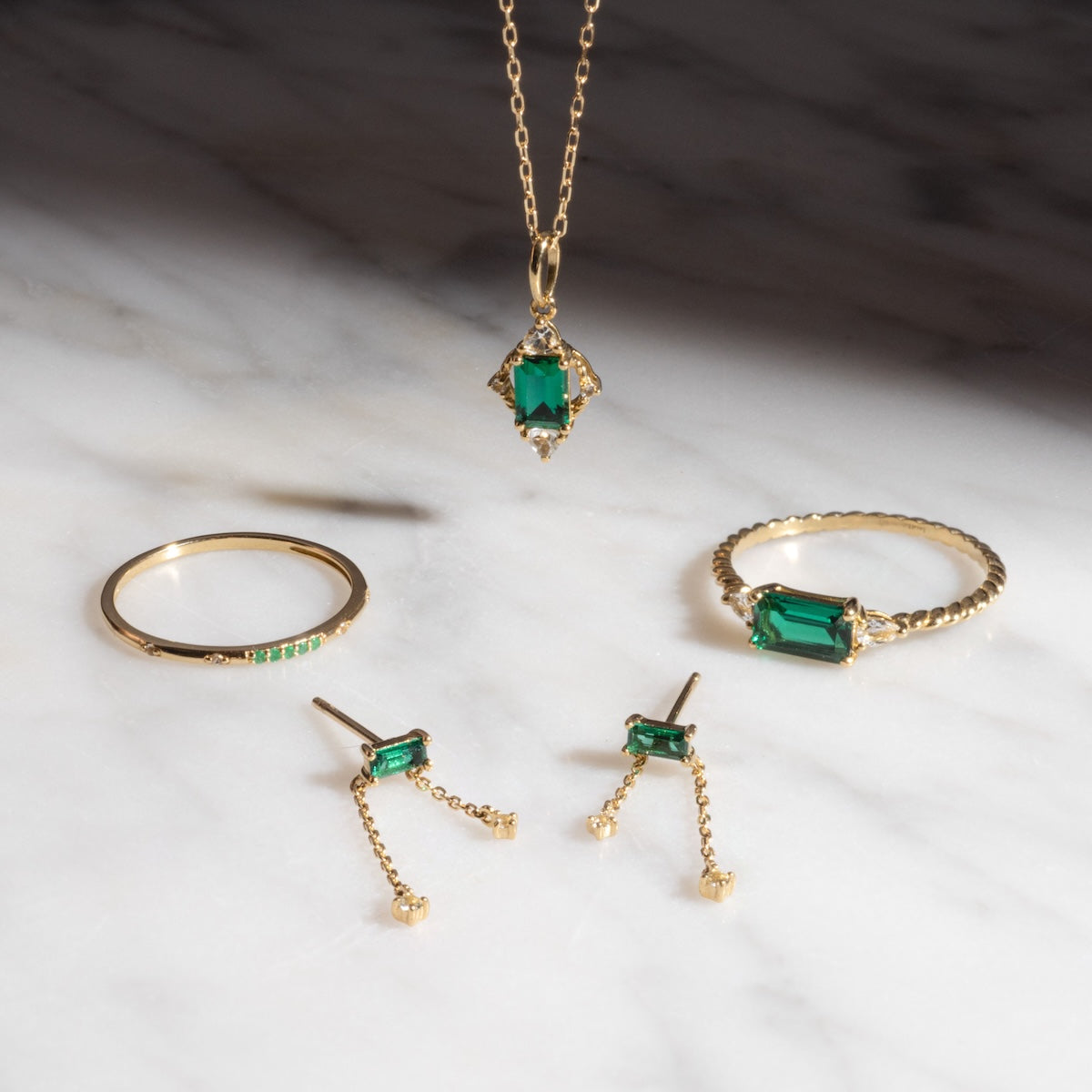 The Solid Gold 2025 May Capsule Delicate Emerald Chain Earrings—perfect for May birthstone lovers—gleam with emerald accents and are elegantly displayed on a white marble surface.