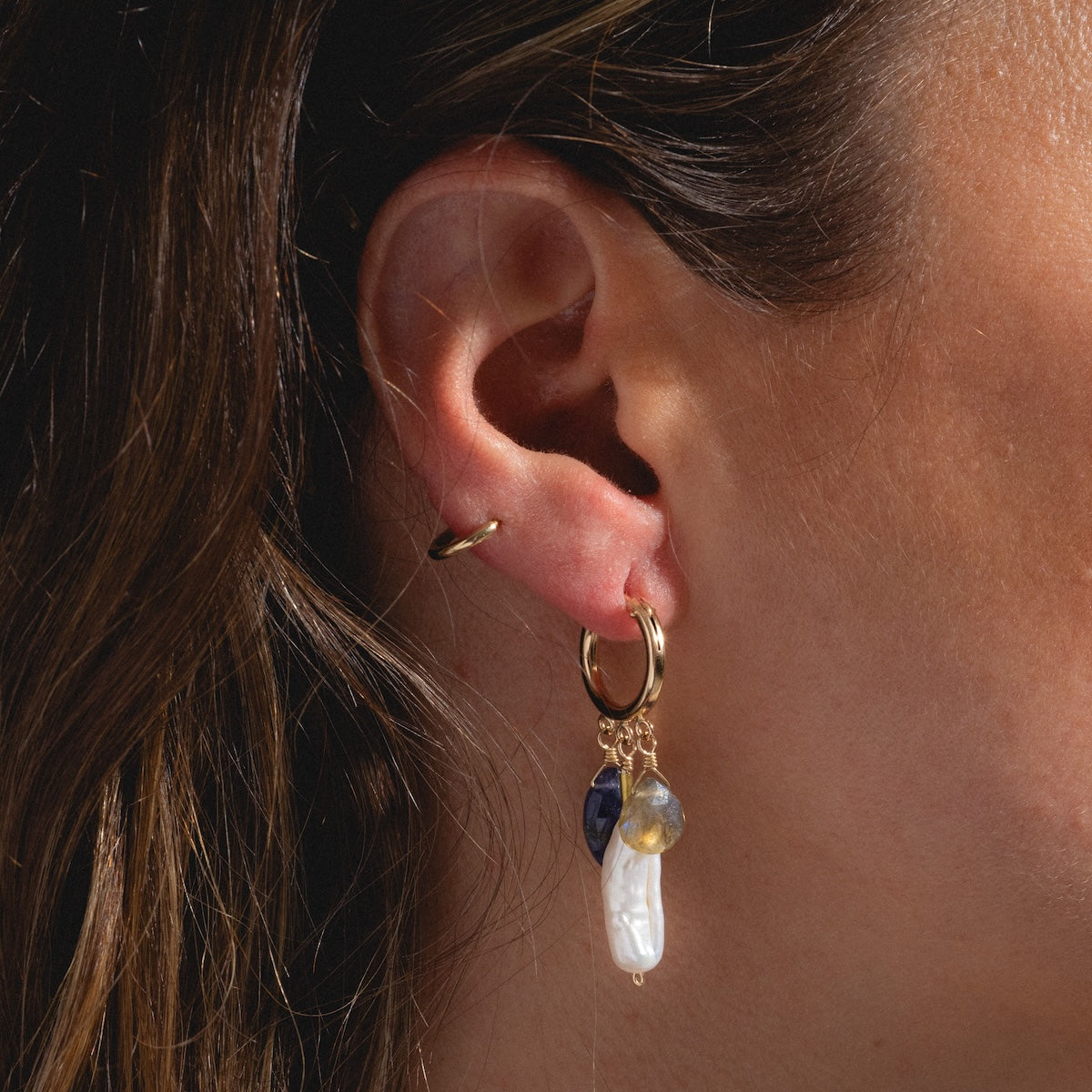 Close-up of a womans ear wearing the Oriana Earrings Sample: one gold hoop and a larger hoop with charms—a baroque freshwater pearl, an iolite stone, and a clear stone. Brown hair is partially visible.