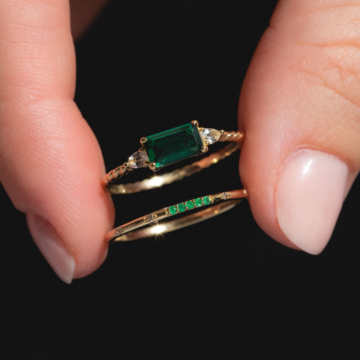 A hand with pale pink nails holds two gold rings: the Solid Gold 2025 May Capsule Classic Emerald Statement Ring, featuring a rectangular green gemstone and side stones, and a slim gold ring with small green stones for birthstone jewelry fans.