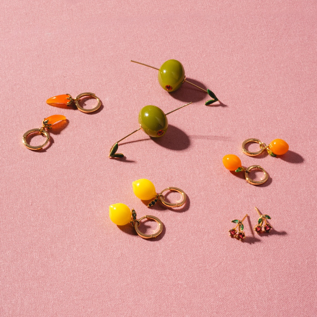 Juicy Cherry Studs are gold-plated earrings in playful cherry shapes, arranged on a pink textured surface. Their colorful, 90’s-inspired design channels fun and whimsy for a vibrant accessory statement.