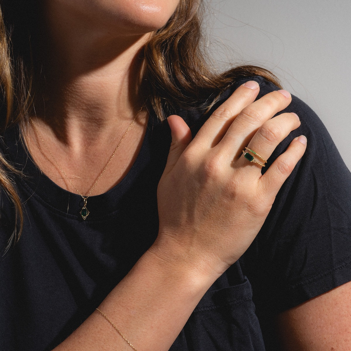 A woman in a black t-shirt models the Solid Gold 2025 May Capsule Classic Emerald Statement Ring, along with gold jewelry like a delicate green birthstone necklace and thin bracelet, against a plain background.