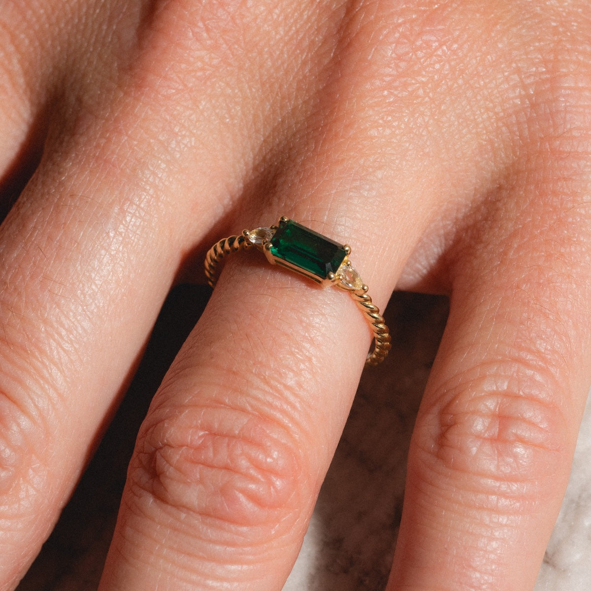 A close-up shows a hand wearing the Solid Gold 2025 May Capsule Classic Emerald Statement Ring, featuring a rectangular green gemstone set in the center and a twisted band, highlighting the natural texture of the skin.