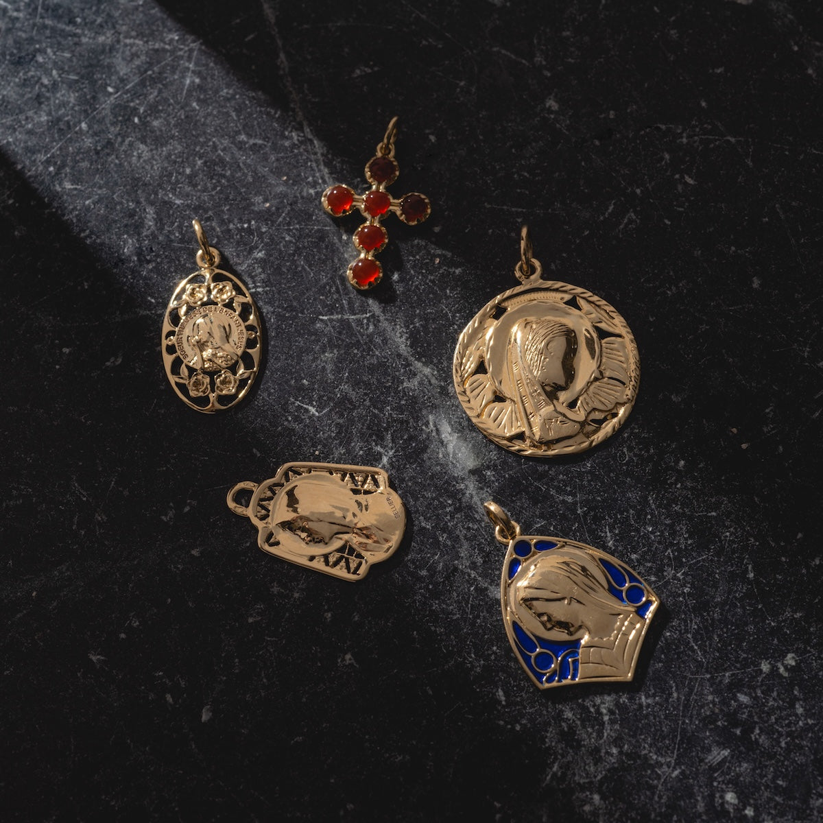 Five gold pendants rest on a dark, textured surface. Four show saintly figures, such as the 14k Vintage Revival Mother Mary Enamel Charm. The fifth is a cross set with red gemstones; light casts gentle shadows over the display.