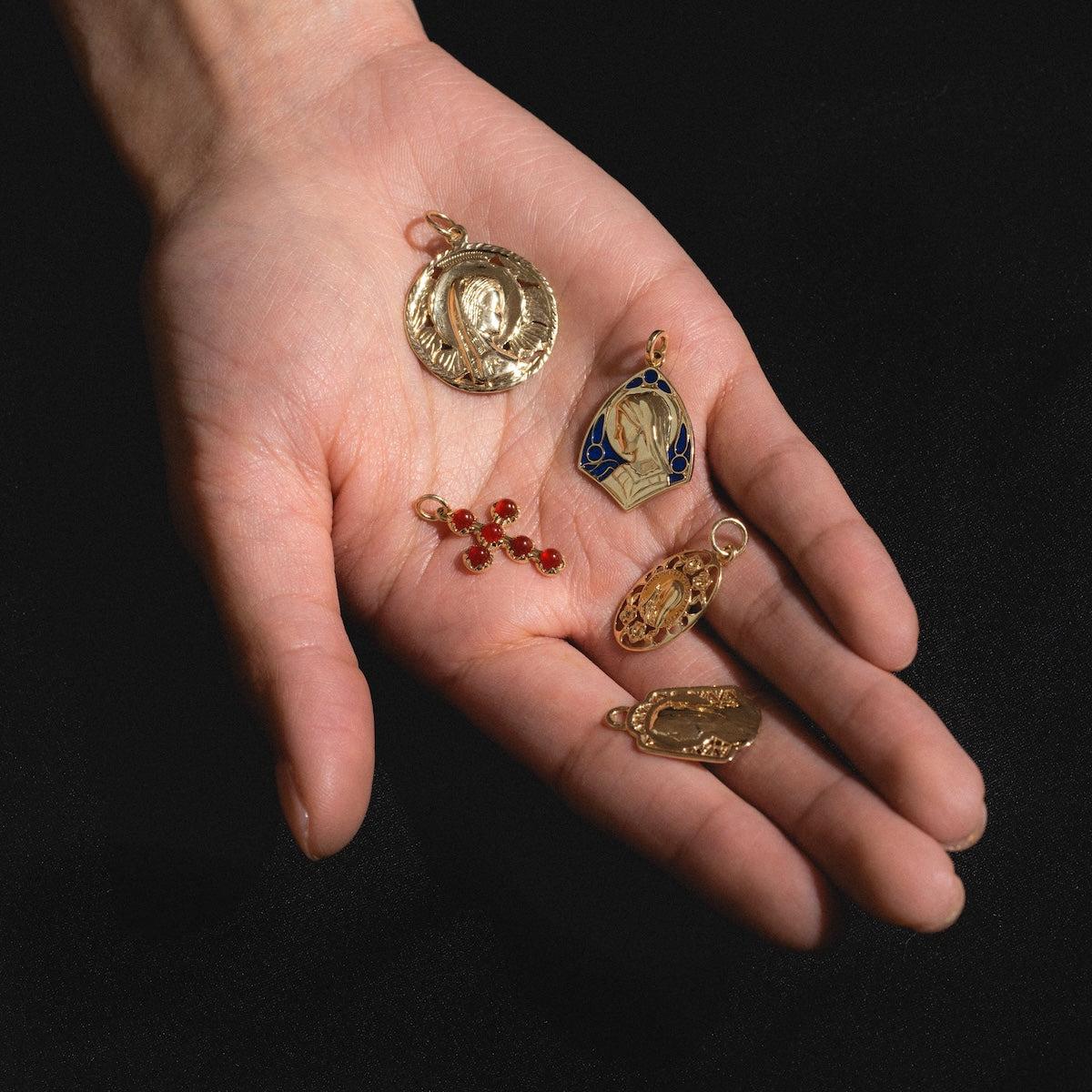 A hand holds five religious pendants, including the 14k Vintage Revival Mother Mary Enamel Charm, saint-engraved vintage jewelry, a gold-and-blue enamel piece, a red gem cross, and ornate gold charms against a black background.