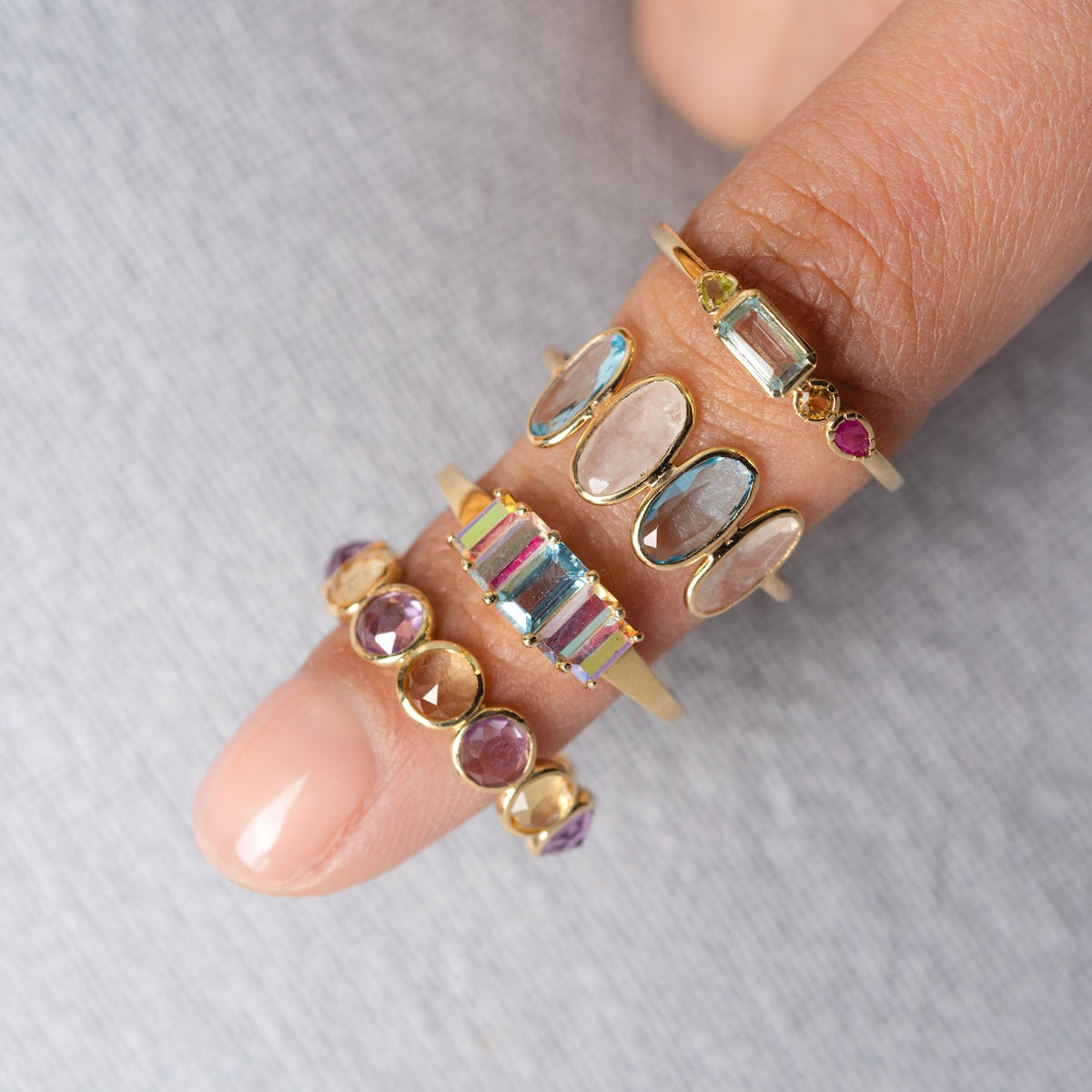 A close-up of a finger wearing the Solid Gold Sunrise Citrine and Alexandrite Band, stacked with solid gold bands featuring colorful gemstones in various shapes and sizes against a light gray textured background.
