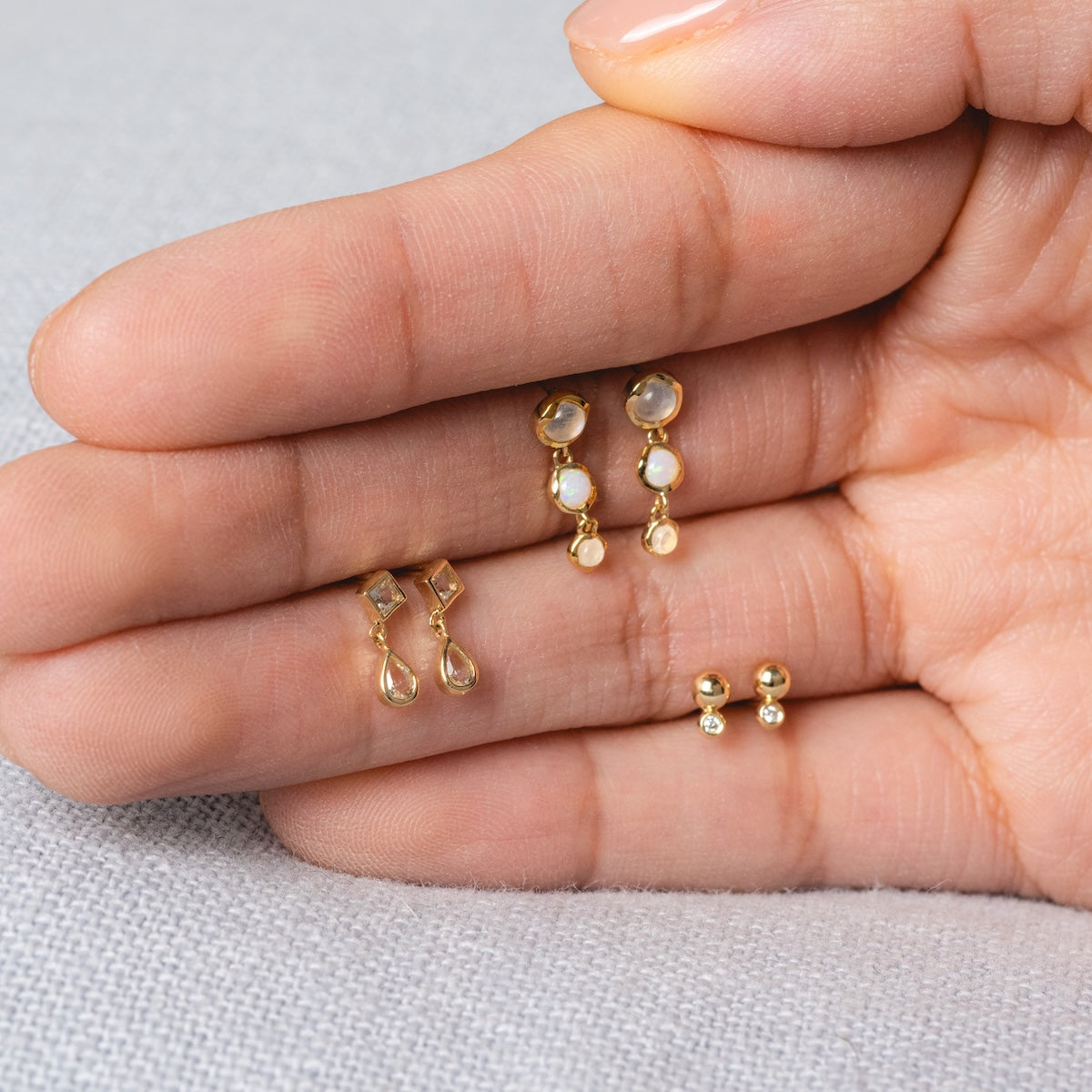 A hand displays three pairs of Solid Gold Horizon Moonstone and Opal Drop Earrings with different designs, resting on the palm against a light gray fabric background.