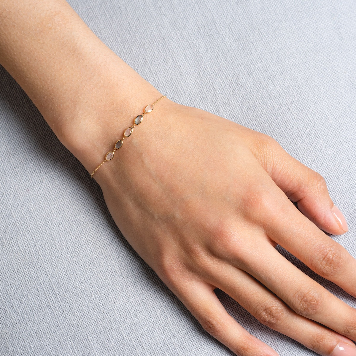 The Solid Gold Daybreak Moonstone and Blue Topaz Bracelet, featuring delicate gemstones, is worn on a wrist and rests on a light gray fabric background.