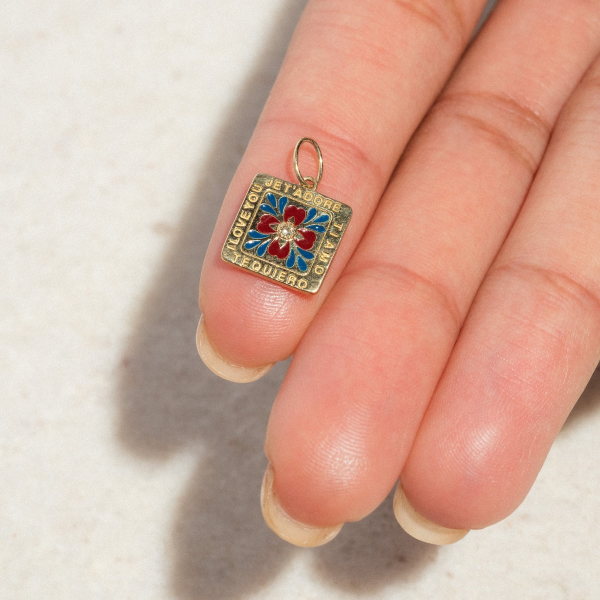 A hand holds the Solid Gold Statement Charm, a small square 9k yellow gold pendant featuring a floral center and I love you engraved in various languages around the edges.