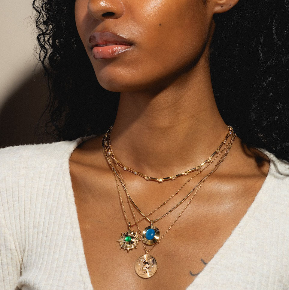 A woman with medium brown skin and curly hair wears a cream sweater and the Saturn Blue Mother of Pearl Pendant Necklace Sample, layered with two other 14k gold plated necklaces featuring a green stone sun and a raised coin pendant.