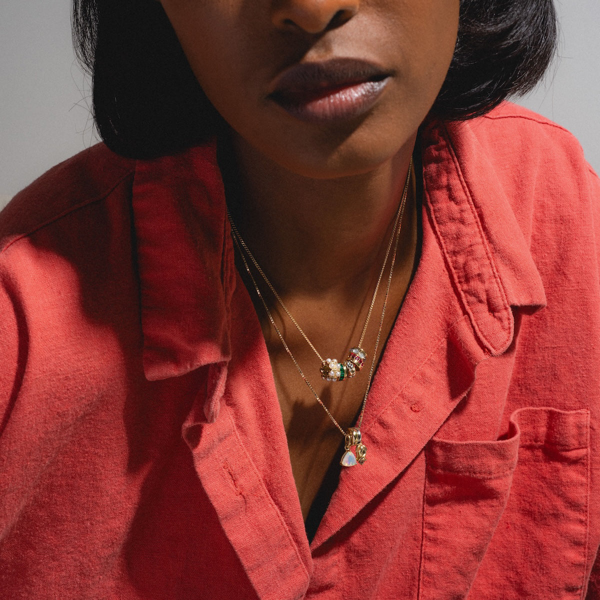 A woman in a coral red shirt wears layered gold necklaces, featuring the Solid Gold Anniversaire Charm with gemstone pendants and floral designs—perfect milestone jewelry—against her dark skin. Her face is partially shown.