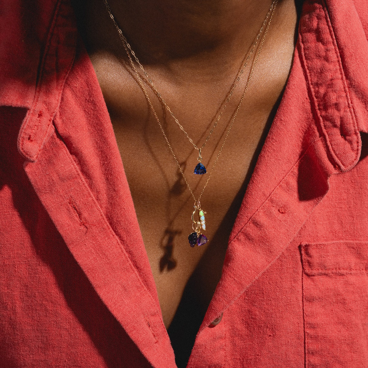 A person wears a softly textured, slightly open red button-up shirt and two gold necklaces—each with a gemstone pendant, including the Family Gold Solid Gold Trillion Birthstone Charm in blue and a purple stone—around their neck.