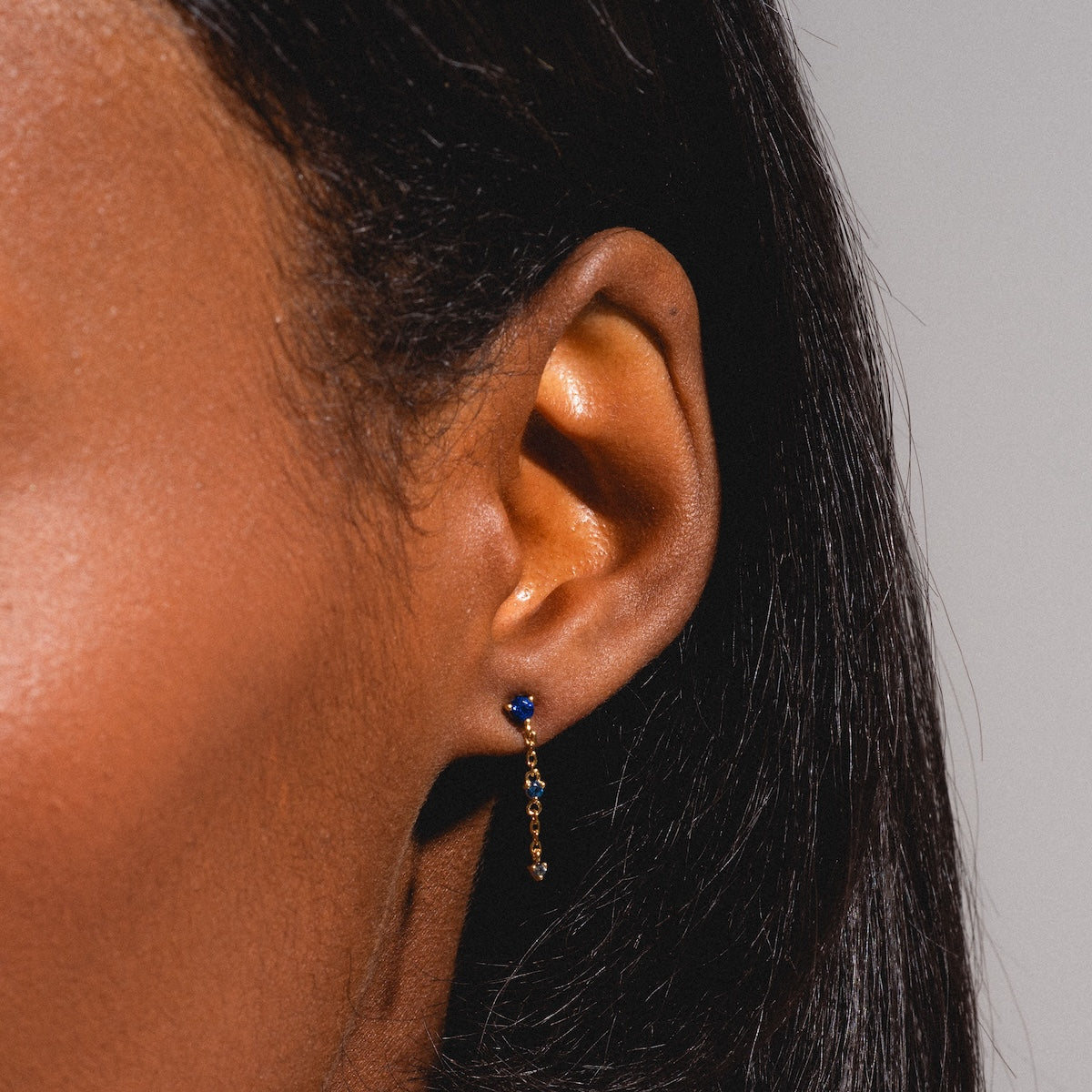 Close-up of a persons ear wearing Celebration Ombre Birthstone Drop Studs—14k gold plated earrings with a blue gemstone and delicate dangling chain. The background is neutral, and their straight, dark hair is partially visible.
