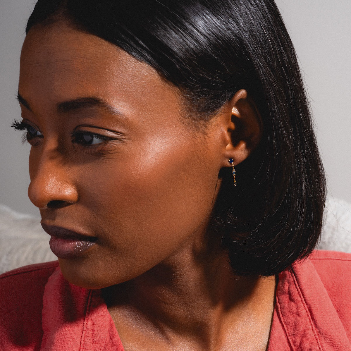 A woman with smooth dark skin and straight shoulder-length hair wears a red top and the Celebration Ombre Birthstone Drop Studs, gazing thoughtfully to the left against a neutral background.