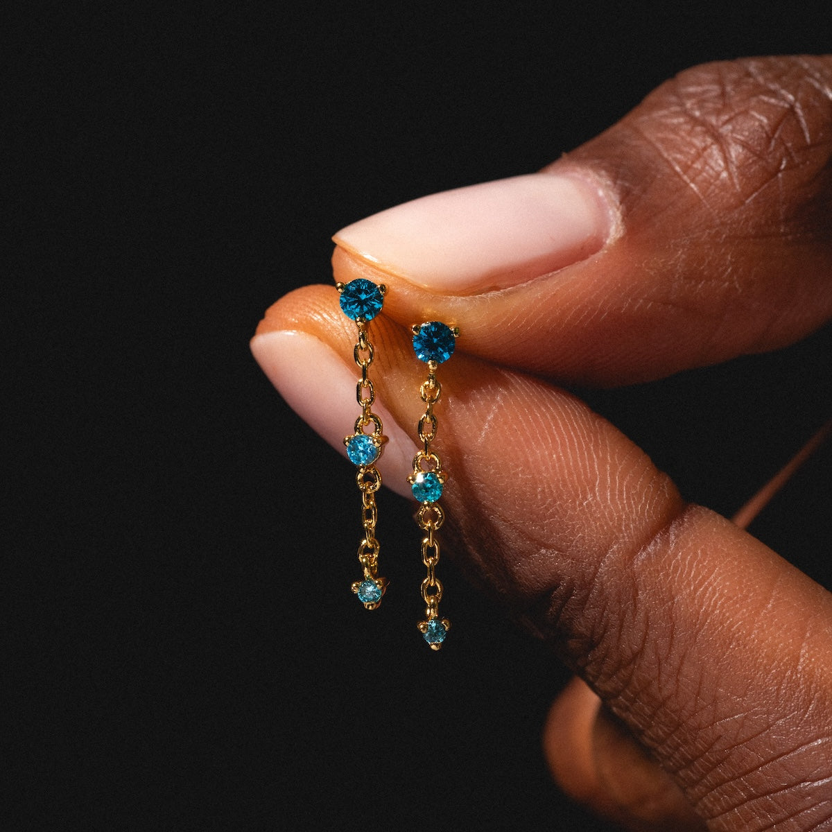A close-up of a hand holding the Celebration Ombre Birthstone Drop Studs, featuring 14k gold plating and small blue gemstones against a dark background, highlighting elegant personalized jewelry.