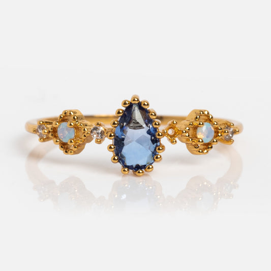 The Something Blue Ring showcases a pear-shaped blue gemstone, flanked by two round opals and clear stones, set in an ornate 18k yellow gold plated band with delicate milgrain detailing.