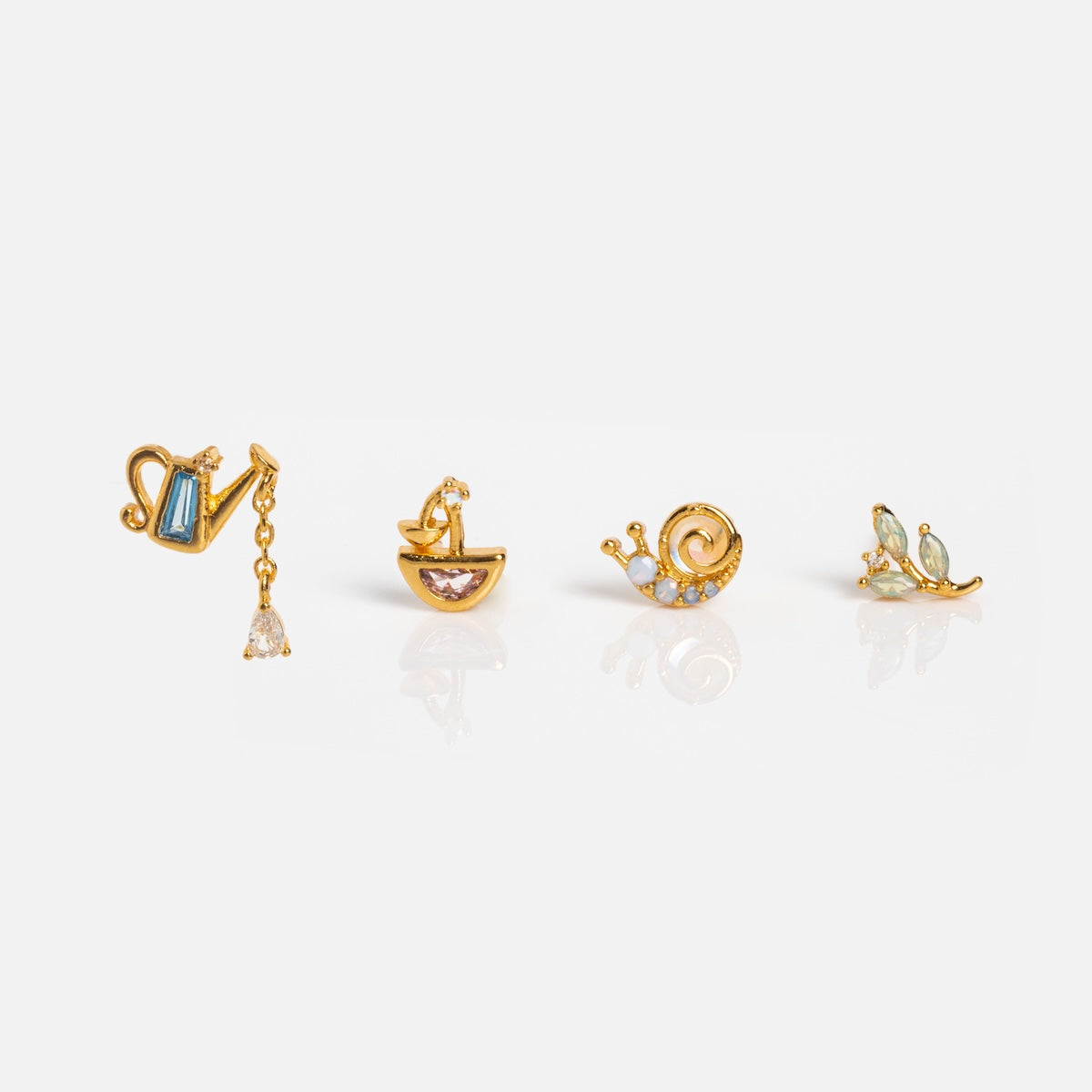 The In the Forest Stud Set includes four small gold plated earrings: a nature-inspired watering can with dangling gem, a sailboat with pink CZ stud, a snail with pearl details, and a branch adorned with three green gems.