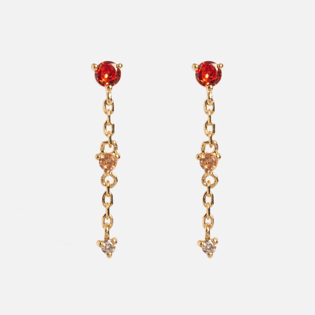 The Celebration Ombre Birthstone Drop Studs are 14k gold-plated earrings with a round red gemstone on top, a smaller gold gem in the center, and a clear gemstone dangling at the bottom.