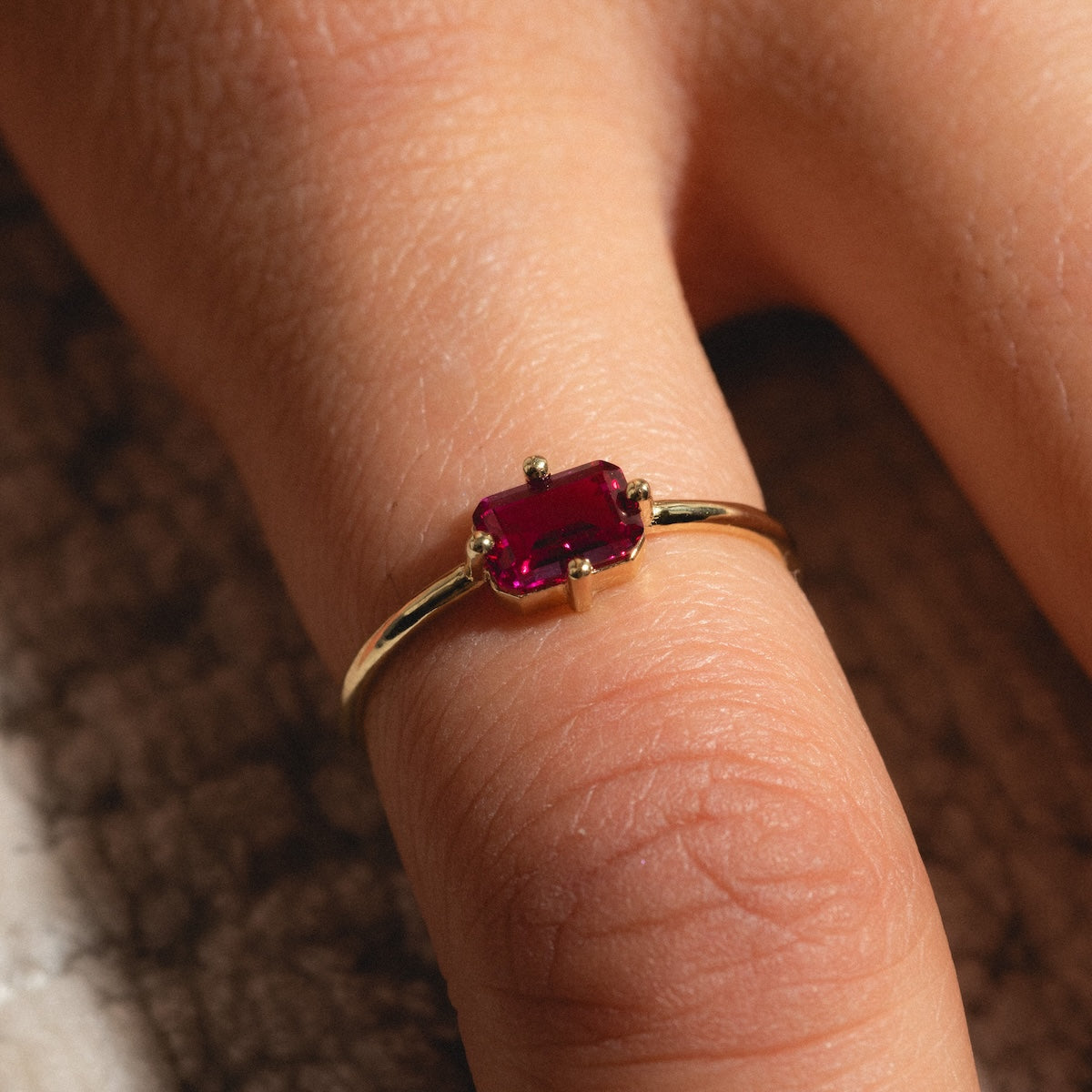 A close-up of a hand wearing the 14k Mod Love Birthstone Ring, crafted in yellow gold with a rectangular deep red baguette gemstone at its center, elegantly displayed on the finger against a softly lit background.