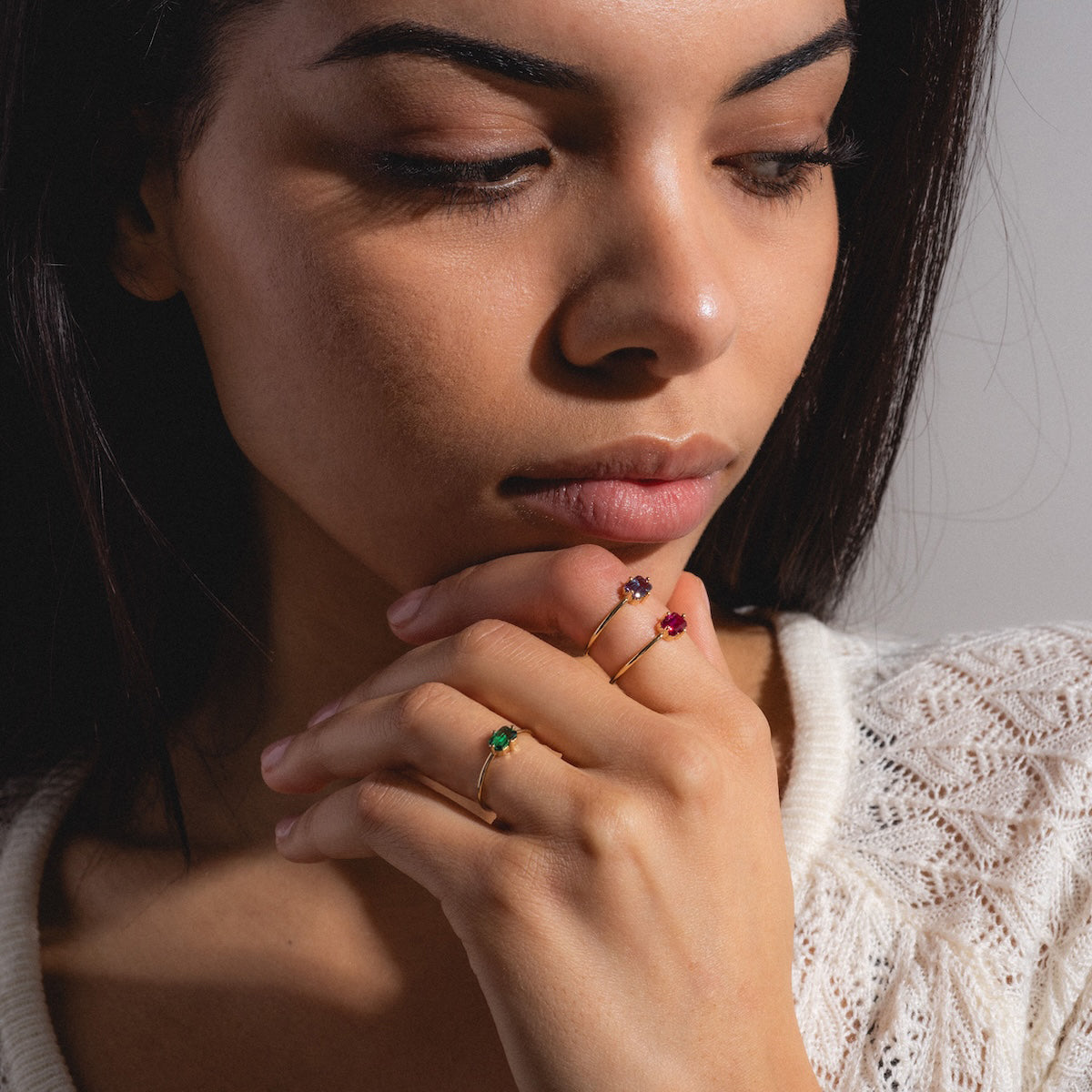A woman with long dark hair, wearing a white textured top, gazes down thoughtfully with her chin on her hand. On her fingers are three 14k Mod Love Birthstone Rings in yellow gold, set with green, purple, and red gemstones.