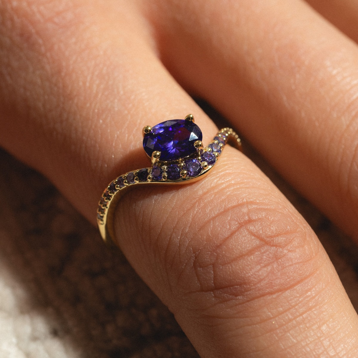 A close-up of a hand wearing the Main Character Floating Ombre Birthstone Ring, featuring a large oval purple gemstone and smaller stones set in a curved band.