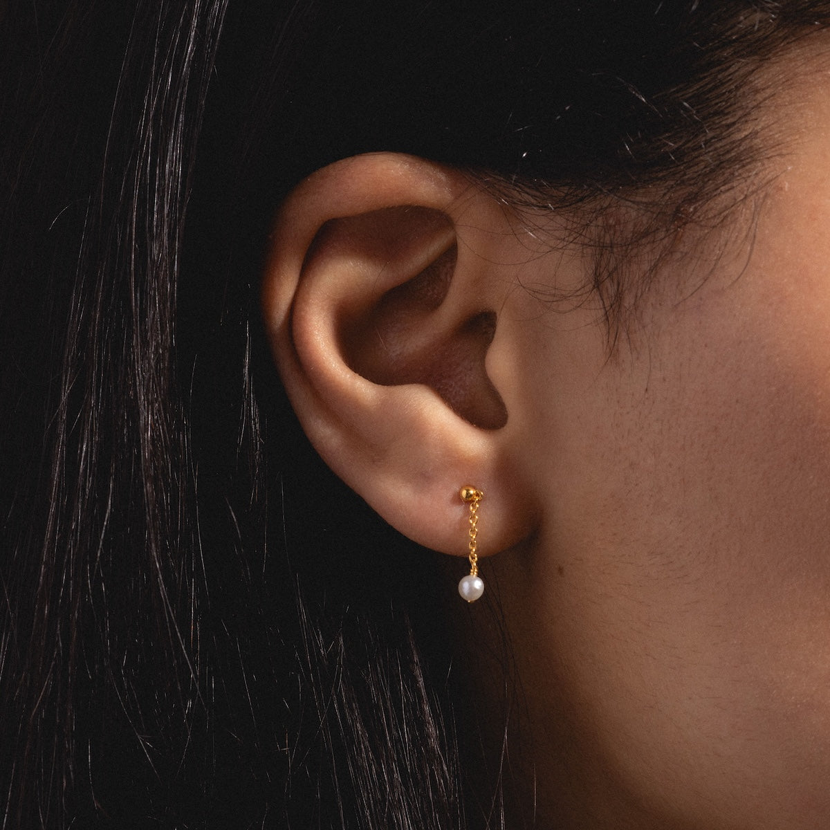 Close-up of a womans ear with long dark hair, wearing the Dainty Pearl Drop Earrings Sample—gold plated drop earrings with a delicate chain and a single white pearl pendant.