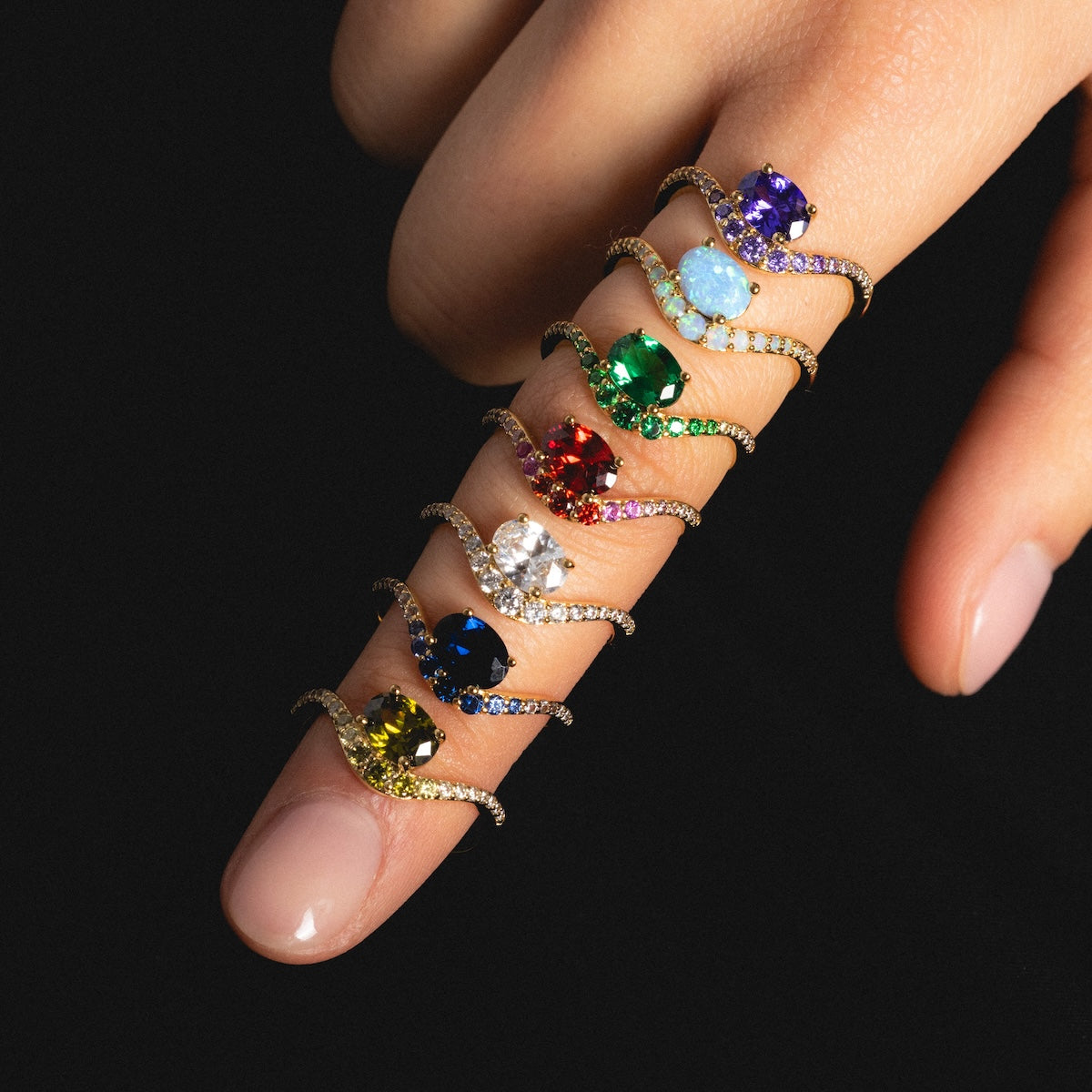 A hand with fingers gently curled displays seven Main Character Floating Ombre Birthstone Rings, each with a unique colored gemstone, arranged from fingertip to base on the index finger against a black background.