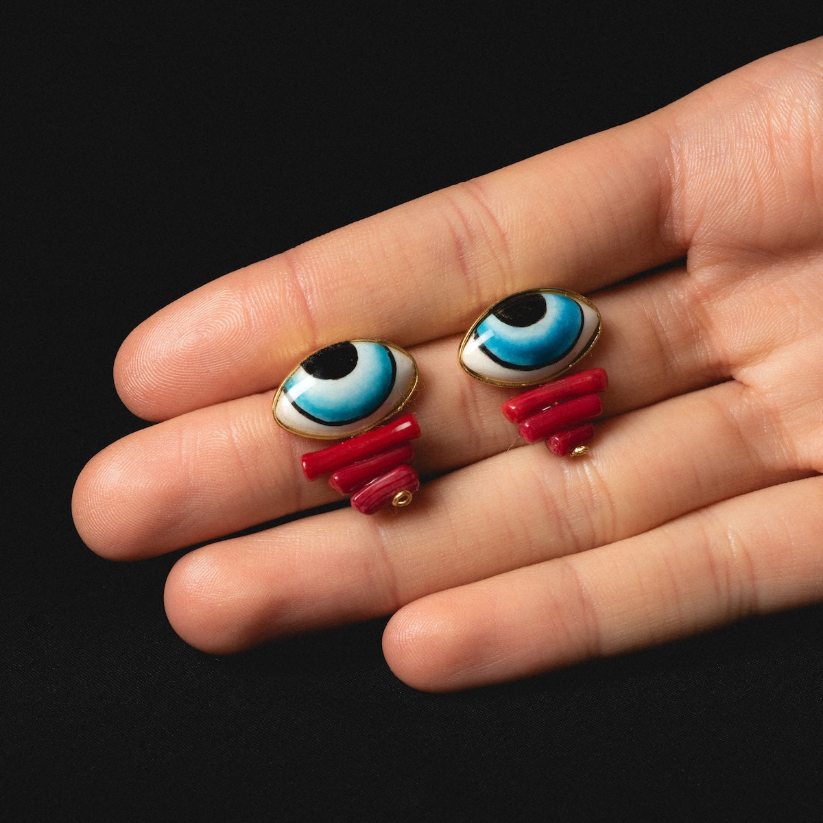 A hand holds Adira Blue Porcelain Evil Eye Stud Earrings, featuring hand-painted blue cartoon eye designs with red bases, displayed against a black background.